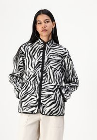Jackets od flisa s uzorkom zebre, visokim ovratnikom, punim prednjim zatvaračem, crnim obrubom i bočnim džepovima. Tkanina ima mekani, teksturirani izgled.