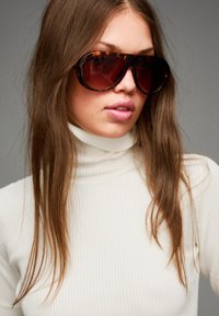 Überdimensionale Sonnenbrille mit einem dunkelbraunen Schildpatt-Rahmen und braunen Verlaufslinsen. Das Modell trägt ein weißes, geripptes Rollkragenoberteil.