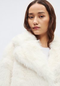 Λευκό παλτό από ψεύτικη γούνα με γιακά σαλίγκαρου, χα�ρακτηριστικό απαλής, αφράτης υφής. Σχεδιασμένο με χαλαρή εφαρμογή και μινιμαλιστική λεπτομέρεια.