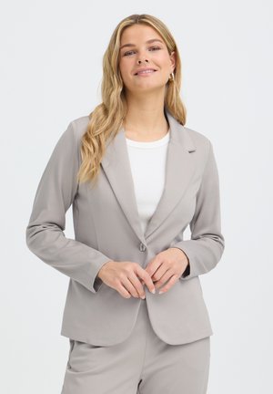 Femme aux longs cheveux blonds portant un blazer gris clair et un pantalon assorti sur un haut blanc, souriante, les mains près du bouton du blazer.