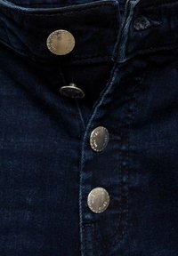 Temno modra denim tkanina z gumbnim zapiranjem na sprednji strani, ki vsebuje pet kovinskih gumbov in diskretne šivane podrobnosti vzdolž zapenjalne vrstice.