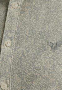 Košulja na dugme u svijetloj bež boji sivo apstraktnim uzorkom, koja sadrži mali izvezeni emblem i okrugle dugmadi. Tkanina izgleda teksturirano.