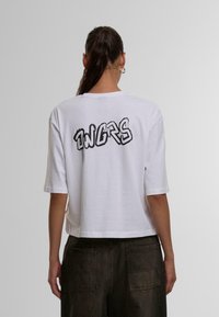 Weißes, kurzes T-Shirt aus Baumwolle. Auf der Rückseite befindet sich der grafische Text "ONGRS" in Schwarz, mit einem leicht abgenutzten Stil. Kurze Ärmel.