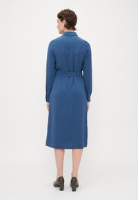 Μπλε φόρεμα με μακρύ μανίκι και γιακά, που έχει ζώνη στη μέση. Λείο υλικό, μέχρι το γόνατο, και κουμπωτές μανσέτες. Φοριέται με παπούτσια με τετράγωνη τακούνι.