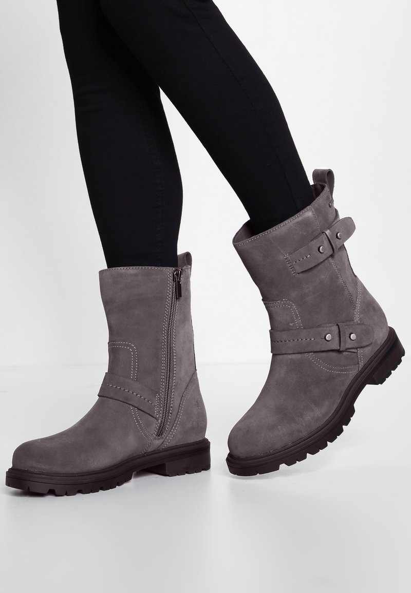 Personne portant des bottines en daim gris avec des fermetures éclair latérales, des sangles et des semelles crantées noires, associées à un pantalon slim noir.