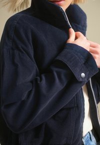 Námořnická kordová bunda s vysokým límcem, zipovým uzavřením a knoflíkovými manžetami. Obsahuje žebrovanou texturu a detail přední kapsy.