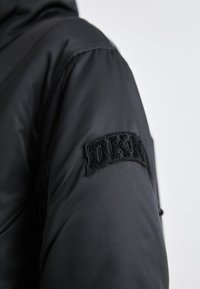 Črn podložen plašč z teksturirano tkanino, ki ima na levem rokavu izrazit črn filcni našitek z napisom "DKNY".