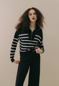 Schwarz-weiß gestreifter Pullover mit Reißverschluss und langen Ärmeln, ausgestattet mit einem goldenen Reißverschluss und einem gerippten Saum, kombiniert mit schwarzen weit geschnittenen Hosen.