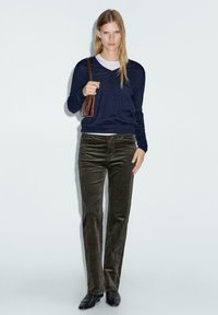 Pullover navy sopra una maglietta grigio chiaro, abbinato a pantaloni in velluto a coste verde oliva. Borsa marrone, scarpe nere, design a gamba dritta, texture liscia.