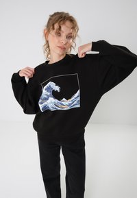 Black sweatshirt with a large wave graphic in blue and white. Features a relaxed fit and ribbed cuffs. Worn with black pants.