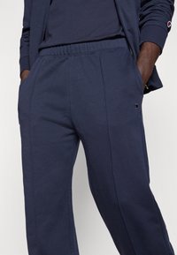 Σκούρο μπλε φόρμα από απαλό ύφασμα, με ελαστική μέση, πλαϊνές τσέπες και διακοσμητικό λογότυπο κοντά στο αριστερό ισχίο.