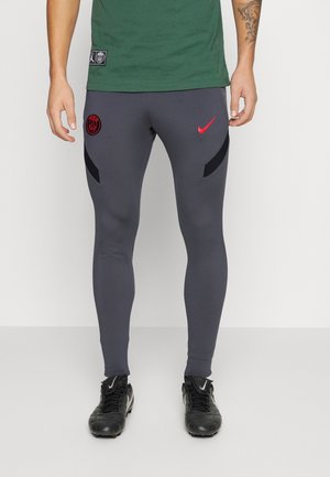 Homme portant un pantalon Nike gris foncé avec des logos rouges et des crampons noirs, debout devant un fond clair uni.