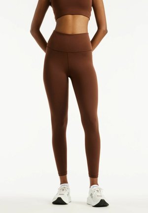 Personne portant des leggings marron taille haute et un crop top assorti, debout les mains dans le dos, chaussée de baskets blanches.
