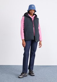 Gilet smanicato di colore verde scuro con fodera nera sopra una felpa rosa chiaro, abbinato a jeans scuri e sneakers grigie su uno sfondo di tappeto blu.