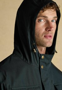 Jachetă cu glugă neagră, cu o textura netedă, dotată cu elemente de metal aurii, nasturi tip buton și o căptușeală rayată vizibilă la guler.