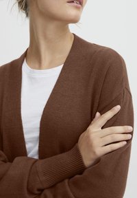 Bruine gebreide cardigan met een sjaalkraag, geribde manchetten en een open voorkant. G gedragen over een wit T-shirt, wat de zachte textuur benadrukt.