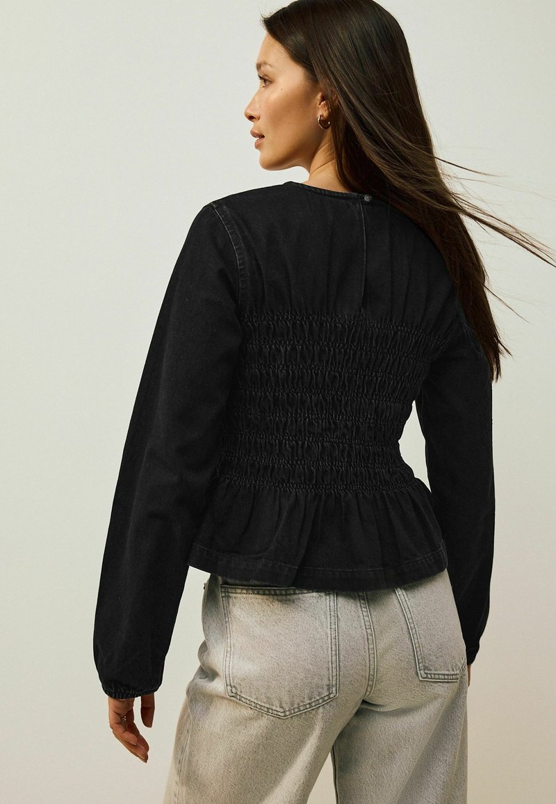 Černá smocked peplum bunda s dlouhými rukávy, s texturovaným zadním dílem a zaobleným výstřihem, v kombinaci se světle šedými kalhotami s vysokým pasem.
