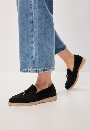 Mocassins en cuir suédé noir avec une semelle en caoutchouc beige, ornés d'un accent doré décoratif, portés avec un jean en denim bleu raccourci.