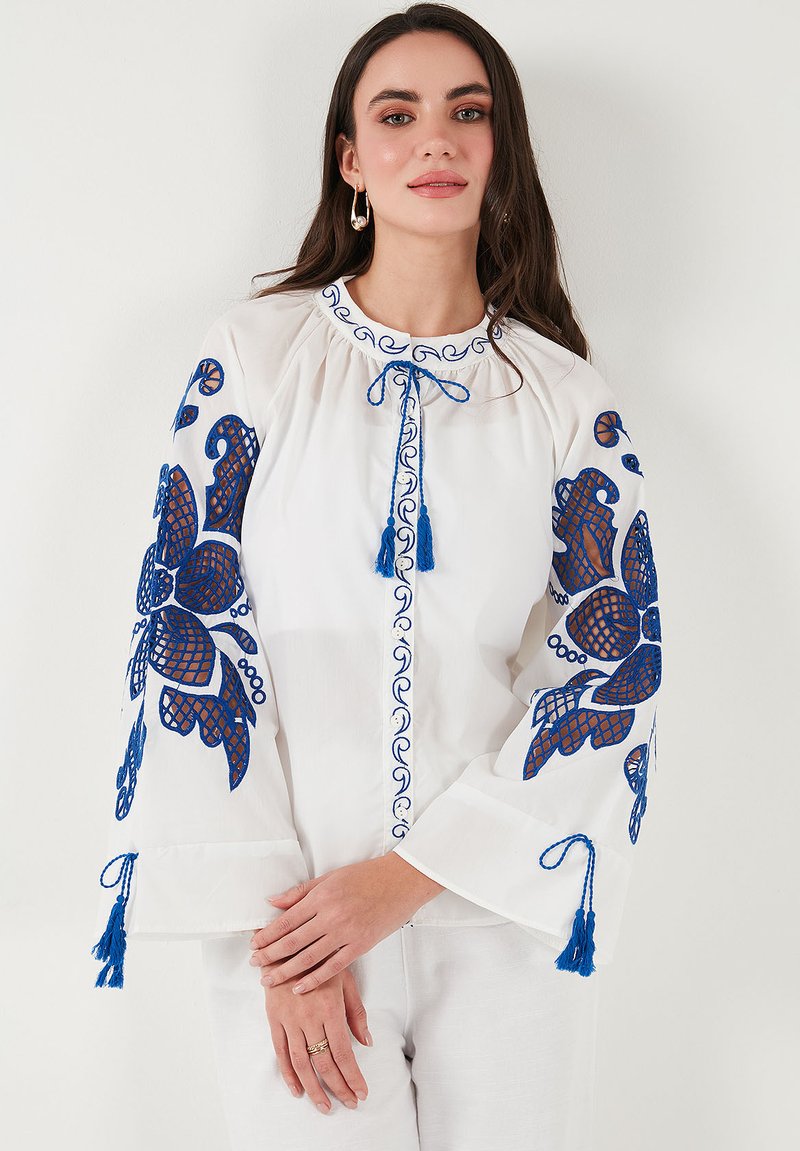 Camicia bianca con ricami floreali blu sulle maniche e sul davanti, caratterizzata da maniche a campana, scollatura rotonda e nappe decorative blu.
