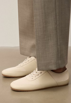 Kremowe buty sznurowane noszone z jasno-brązowymi, dopasowanymi spodniami na neutralnej podłodze, ukazujące szczegóły butów z boku i od góry.