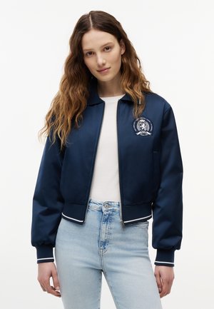 Ung kvinde med langt, bølget hår iført en marineblå bomberjakke med emblem, hvid skjorte og lyseblå jeans, stående mod en hvid baggrund.