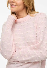 Femme portant un pull rose clair à petits pompons blancs, souriant doucement, avec la main près du visage.