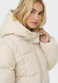 Chaqueta de plumas color crema con diseño acolchado, gran capucha y cierre de botón oculto. Textura suave y ligero brillo.