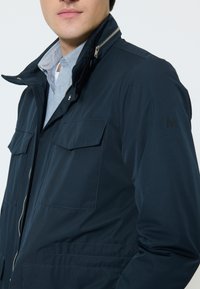 Man wearing a navy blue jacket with zipped collar and flap pockets over a light blue button-up shirt against a plain background.