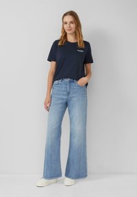 T-shirt bleu marine avec logo graphique, associé à un jean large bleu clair. Le modèle porte des baskets plateforme blanches texturées, se tenant devant un fond neutre.