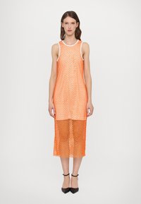 Robe en maille orange avec un design sans manches, présentant un motif géométrique et une longueur de genoux. Portée avec des chaussures noires à bout pointu.