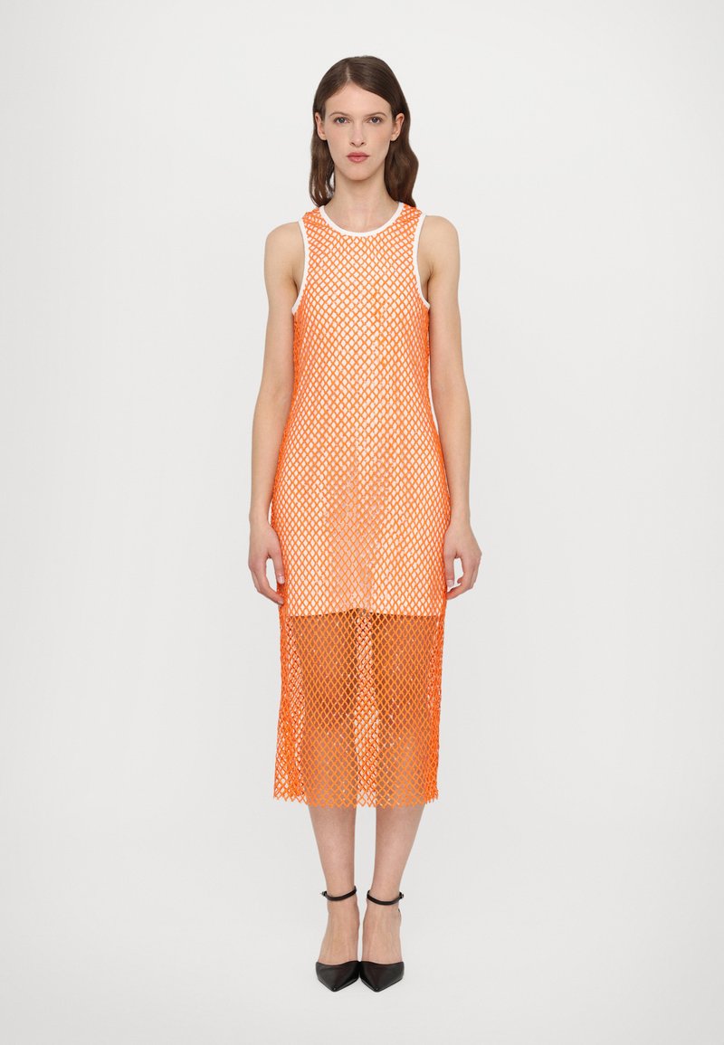 Robe en maille orange avec un design sans manches, présentant un motif géométrique et une longueur de genoux. Portée avec des chaussures noires à bout pointu.