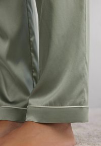 Lichtgroene satijnen pyjamabroek met een soepele textuur, voorzien van witte piping langs de zoom. De stof valt zachtjes.
