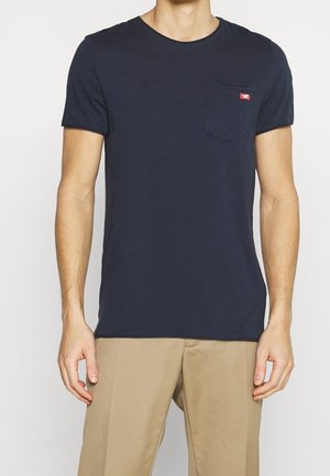 Mann trägt ein dunkelblaues Kurzarm-T-Shirt mit Brusttasche und beiger Hose, steht vor einem einfarbigen hellen Hintergrund.