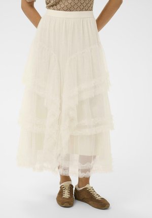 Žena nosi vrstvnatú bielu tylovú midi sukňu v kombinácii s hnedými semišovými teniskami na šnurovanie, stojaca pred jednoduchým pozadím.