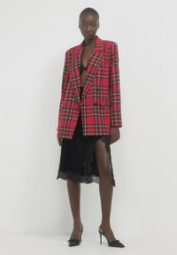 Blazer a quadri rossi con bottoni doppi, abbinato a un vestito in pizzo nero. Le scarpe sono tacchi neri. Lo sfondo è bianco puro.