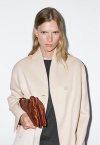 Crème wollen jas met één knoop, grijze top en een bruin leren clutch die in de hand wordt gehouden. Glad textuur en minimalistisch design.