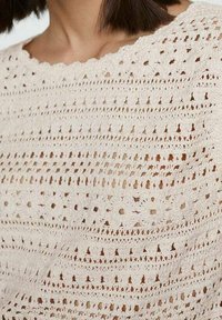 Kremowy top na szydełku z otwartym splotem, w różnych wzorach, z falbaniastym dekoltem i teksturowanym materiałem w luźnym fasonie.