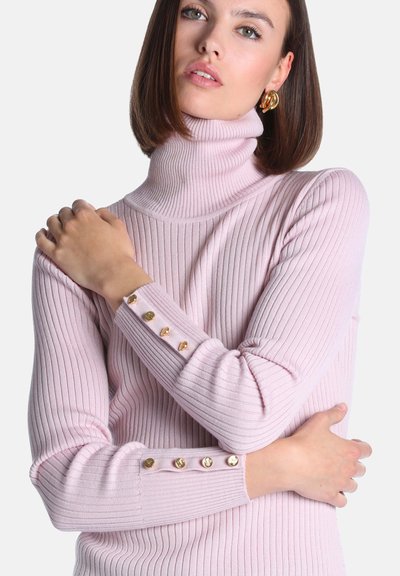 Femme aux cheveux bruns raides portant un pull à col roulé côtelé rose clair avec des boutons dorés sur les manches, les bras croisés.