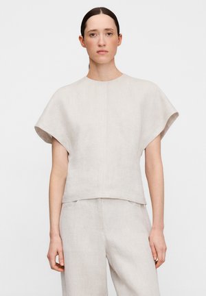 Vrouw met donker haar naar achteren gebonden, gekleed in een losse, korte mouw, lichtbeige top en bijpassende broek tegen een effen achtergrond.