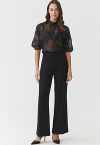 Bluză neagră transparentă cu model floral și mâneci scurte evazate, asociată cu pantaloni negri cu talie înaltă. Modelul poartă pantofi negri cu vârful ascuțit.