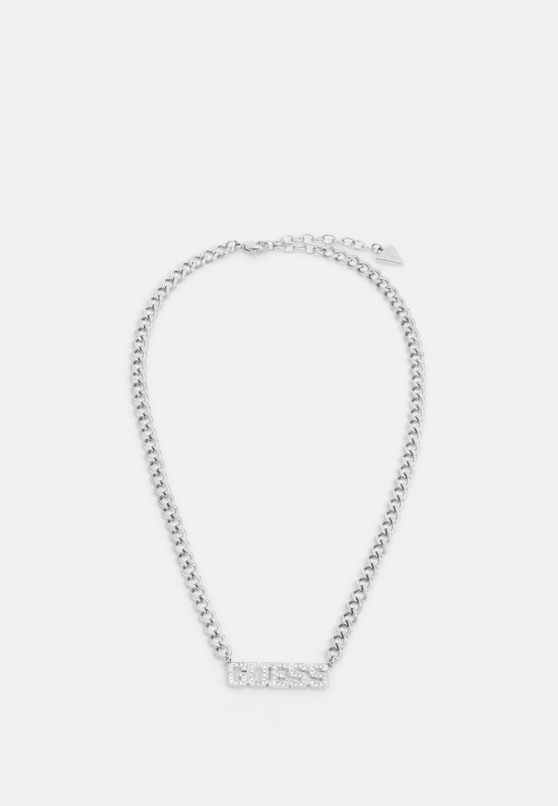 Collier chaîne en argent avec un pendentif rectangulaire affichant "GUESS" en petits cristaux et une petite étiquette triangulaire près du fermoir.