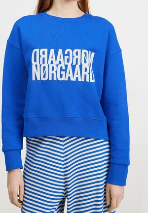 Osoba ubrana w jasnoniebieską bluzę z białym, zniekształconym napisem "NØRGAARD", w połączeniu z niebiesko-białymi, poziomo paskowanymi spodniami.