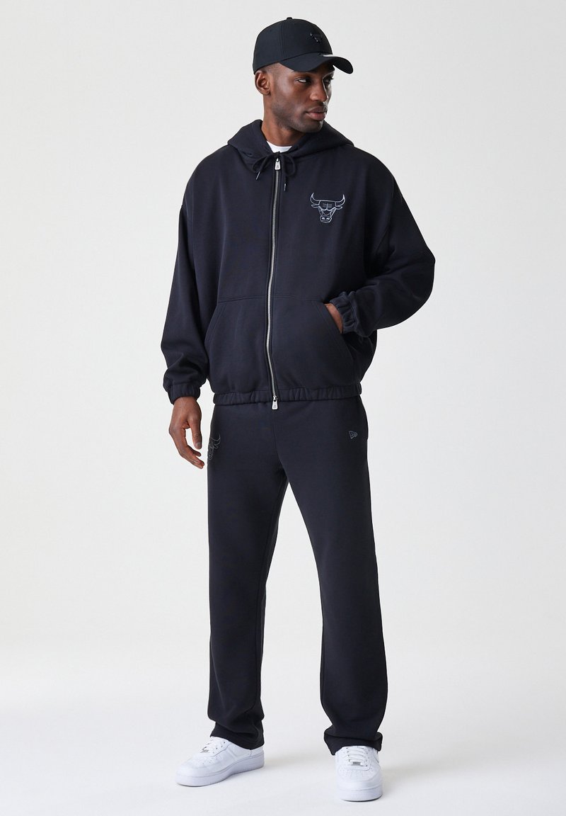 Sudadera negra con cremallera y logo en el pecho, combinada con pantalones de chándal a juego. La tela parece suave; se lleva con zapatillas blancas y una gorra.