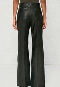 Kobieta w czarnych, skórzanych, szerokich spodniach o wysokim stanie z tylnymi kieszeniami, stojąca na jednolitym jasnym tle.