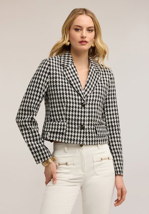 Kvinde iført en sort- og hvid hundetandsmønstret blazer med to knapper og hvide højtaljede bukser med frontlommer og gulddetaljer.