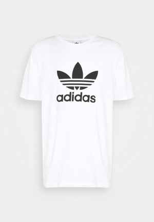 Mężczyzna ubrany w zieloną i czarną koszulę w kratę na białej koszulce z logo Adidas, czarne podwinięte dżinsy i czarne sneakersy Adidas z białymi paskami.