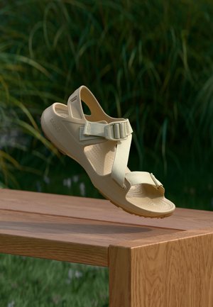 Sandale beige avec des brides réglables flottant au-dessus d’un banc en bois en plein air avec un feuillage vert en arrière-plan.