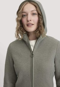 Frau mit lockigem, blondem Haar, die eine hellgraue Zip-Jacke mit Kapuze über einem weißen Oberteil trägt, die frontal vor einem schlichten Hintergrund steht.