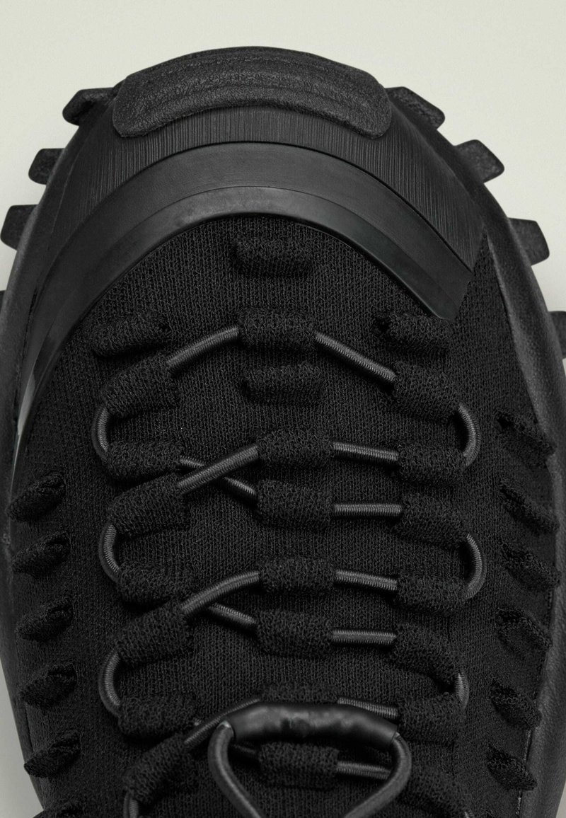 Schwarzer Sportschuh mit einer strukturierten Oberseite, elastischem Schnürsystem und robustem Außensohlenprofil. Auffällige Gummi-Akzenten und ein elegantes, stromlinienförmiges Design.