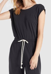 Zwarte jumpsuit met korte mouwen, elastische taille en wittekoord met kwastjes. Zacht materiaal met een ontspannen pasvorm en ronde hals.